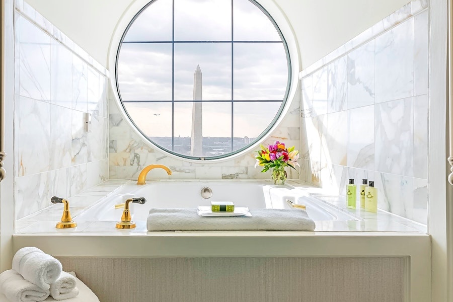 A luxurious bathroom with a marble bathtub and golden fixtures, featuring a round window with a view of the Washington Monument. The bathtub has towels, flowers, and toiletries arranged neatly.