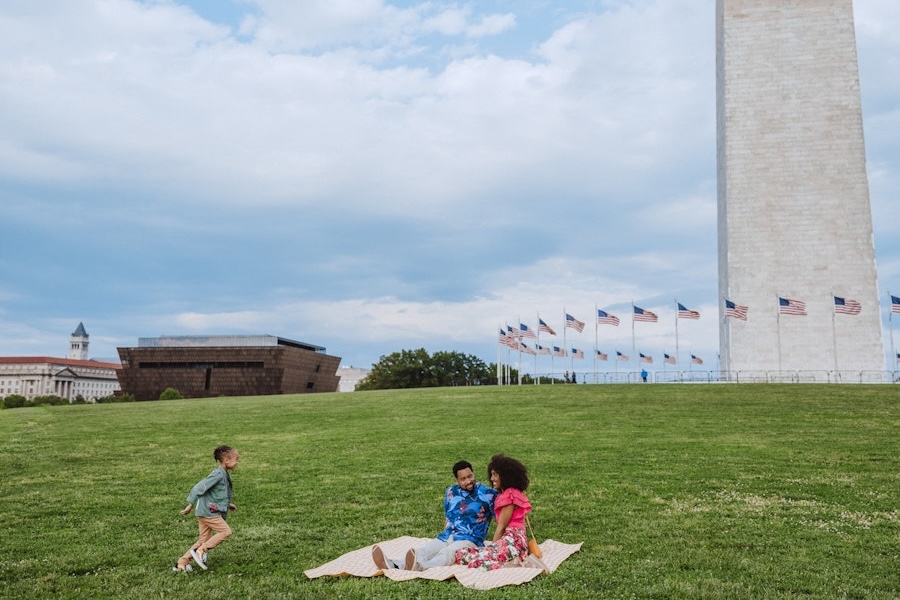 Family enjoying a picnic on the National Mall, with a young boy running towards them. The Washington Monument and the National Museum of African American History and Culture are visible in the background.