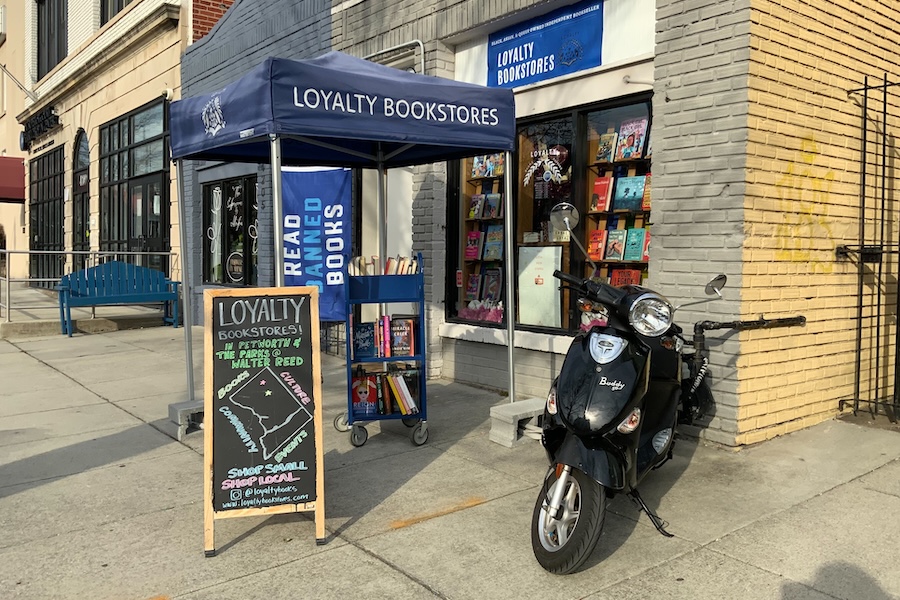 The exterior of Loyalty Bookstores with a blue canopy, book cart, chalkboard sign, and scooter parked out front.