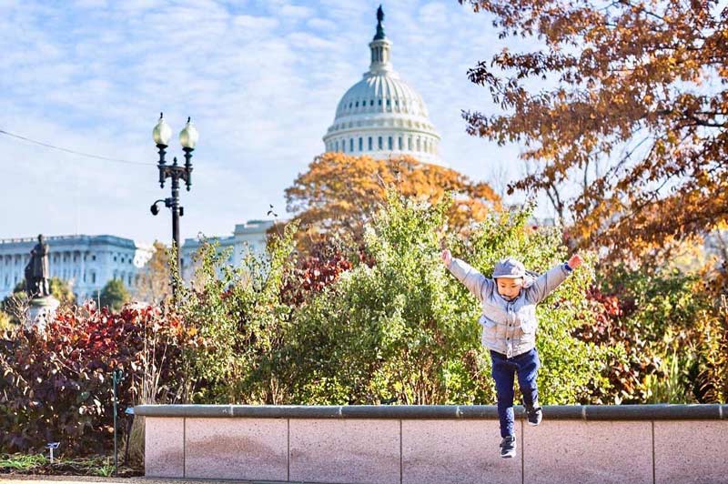 @chasingkaiphoto - Child jumping in front of the U.S. Capitol building surrounded by fall foliage - Fall in Washington, DC