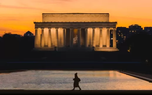A silhouette of a person walking along the reflecting pool is set against the illuminated Lincoln Memorial at sunset, with a warm orange sky and city buildings in the background.
