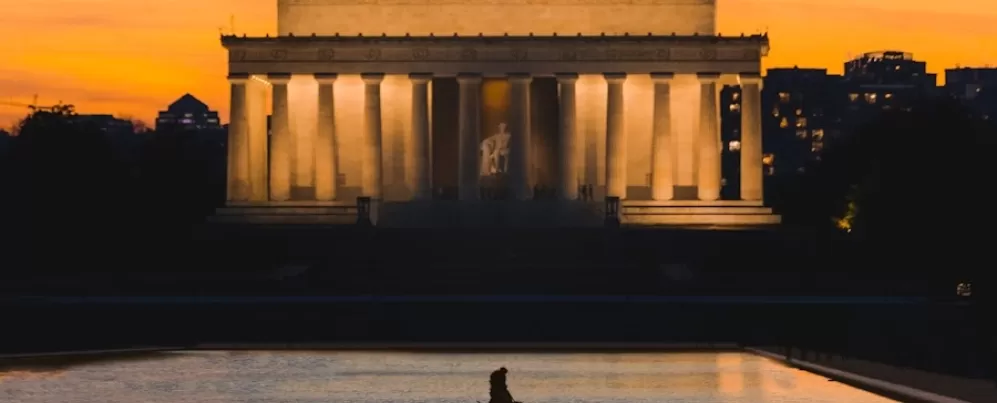 A silhouette of a person walking along the reflecting pool is set against the illuminated Lincoln Memorial at sunset, with a warm orange sky and city buildings in the background.
